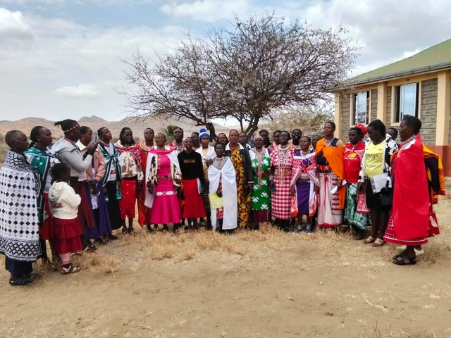 About thirty women in bright shukas and dresses standing together in front of a tree and a training building on dry ground.