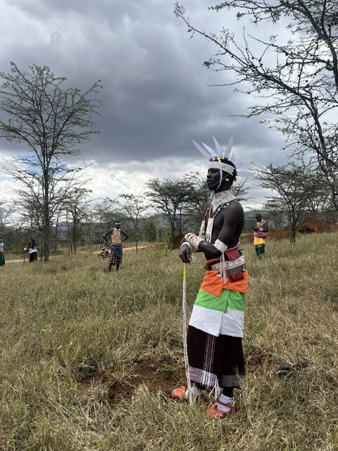 A Moran participant stands in open grassland under a stormy sky, with other participants behind him among acacia trees.