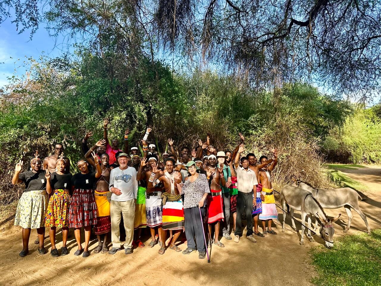 A large group of LAMP participants, many in traditional beaded dress, wave at the camera with Guy Pfeffermann under acacia trees, two donkeys beside them.