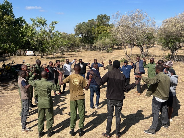 A large group of people, many in green uniform shirts, standing in a circle outdoors in a dry clearing with acacia trees behind them.