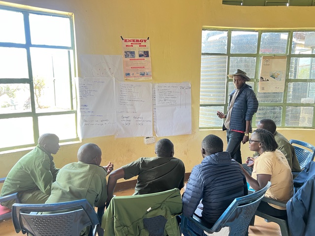 A facilitator in a blue jacket and hat standing at the front of a classroom while participants in green shirts sit in a circle of blue chairs.