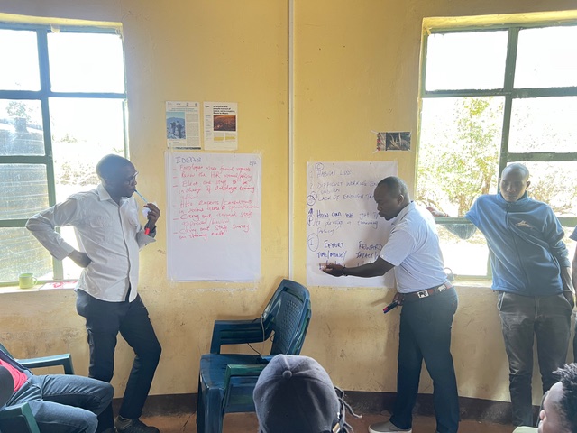 Three men standing in a yellow-walled classroom beside flip-chart sheets covered in handwritten notes.